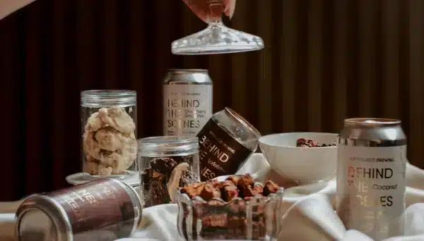 Latas de cerveja artesanal Joy Project Brewing em composição com petiscos variados, sobre tecido branco, com mão segurando tampa de vidro