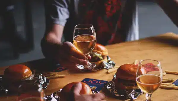 Mesa com hambúrgueres, batatas fritas e copos de chope artesanal na Joy Project Brewing, com mãos segurando copos em um brinde